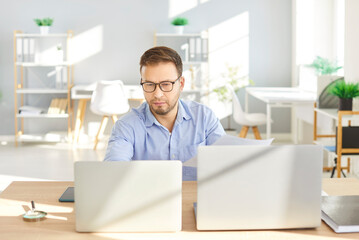 Confident young attractive business man in formal shirt and glasses looking seriously at computer monitor screen working at the desk on his workplace and typing on laptop in office.