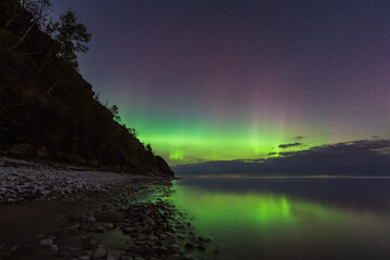 Naklejka premium Stunning green and purple Aurora Borealis (Northern Lights) reflecting in calm water on the rocky coast of Estonia.