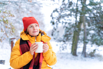 Happy woman wearing yellow jacket and red hat holds hot drink in white mug while sitting in snowy landscape, reflecting peacefully amid nature’s winter beauty. Connection to nature and quiet solitude