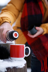 Close-up of senior woman hands pouring hot tea from vacuum flask into red mug outdoors in snowy winter setting. Outdoor comfort and warmth in winter. Simple pleasures as warming drink in cold weather