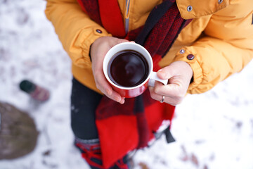 Top view of woman with red scarf, hands holding hot drink in red mug while sitting outdoors in snow and enjoying relaxing winter moment. Cozy solitude, winter relaxation and simple living in nature.