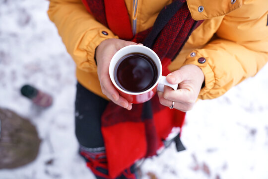 Top view of woman with red scarf, hands holding hot drink in red mug while sitting outdoors in snow and enjoying relaxing winter moment. Cozy solitude, winter relaxation and simple living in nature. - Powered by Adobe