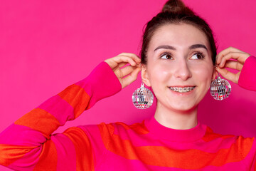 Close-up of a smiling girl in a pink striped sweater with Christmas balls on a pink background. A joyful teenager with braces is laughing and having fun with disco balls.