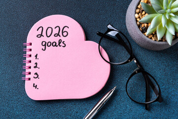 New year resolutions 2026 on desk. 2026 goals list with pink notebook, plant on blue background....