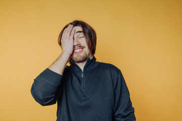 Man with long hair and beard smiling with hand on face against yellow background.