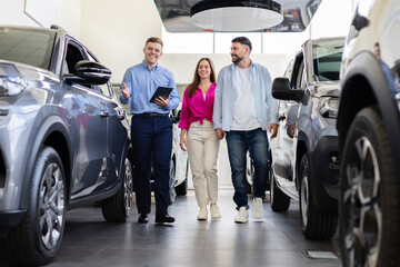 Young couple walking with car salesman inside modern dealership. Smiling customers exploring vehicles, showing trust and enthusiasm in an automotive showroom