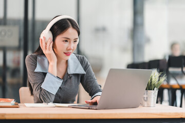 Calm, focused asian woman professional working and listening with headphone on laptop for e learning in bright modern office setting