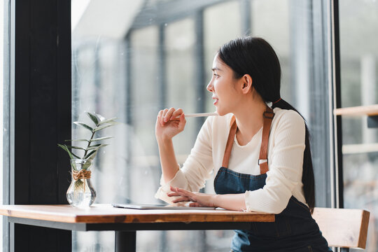 Pensive asian woman cafe owner sitting at table thinking and looking out window. small business entrepreneur having thoughtful break moment - Powered by Adobe