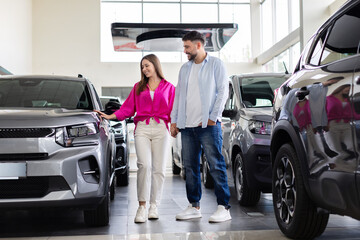 Couple standing together in dealership, woman touching car surface while man watches. Smiling people examining new vehicle before purchase decision
