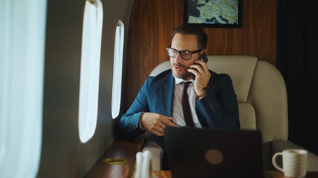 Successful middle-aged businessman in a suit having a business phone conversation while traveling in a luxury private jet, gesturing and smiling during the call with a laptop on the table