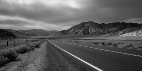 Dramatic monochrome landscape featuring a long road leading towards distant mountains beneath an overcast sky creating a moody travel scene