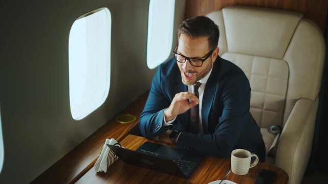Confident middle-aged executive having a business video conference on a laptop computer while traveling in a luxurious private airplane, sitting in a comfortable first-class seat