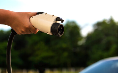 A female hand holds a charging cable of an electric car against the background of the sky and nature. The driver holds a cable for charging an electric vehicle. Clean alternative energy concept.