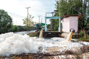 Murky foam piles created by floating fast from flow of polluted water from a large water pump to accelerate drainage after rain at irrigation canal gate along village road in rural Thailand.