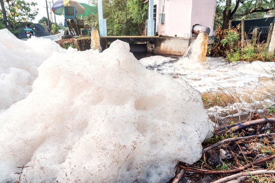 Murky foam piles created by floating fast from flow of polluted water from a large water pump to accelerate drainage after rain at irrigation canal gate along village road in rural Thailand.