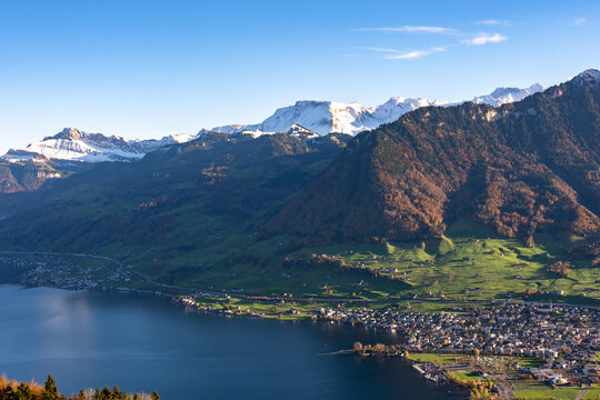 A high-angle view captures a densely settled lakeside town nestled between dark blue water and steep, sun-drenched alpine slopes with snow-capped peaks against a clear sky.