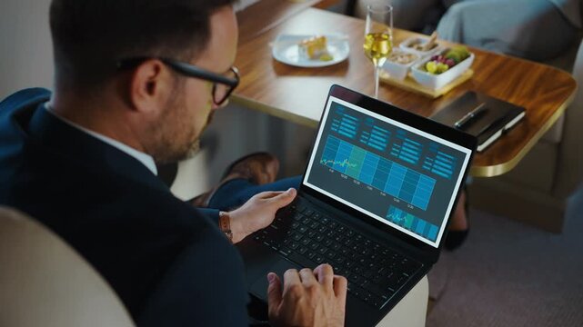 Confident financial analyst working on a laptop, checking stock market charts and investment statistics during a comfortable first-class flight while enjoying snacks and champagne