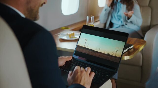 Focused businessman typing on his laptop computer while traveling in a luxury private jet, while his colleague in the background is talking on the phone and enjoying champagne and snacks