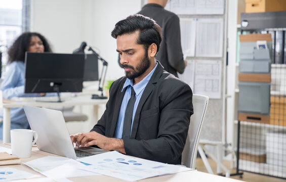 Serious Indian businessman working on laptop in modern office, focused on professional tasks, career development, and corporate productivity.