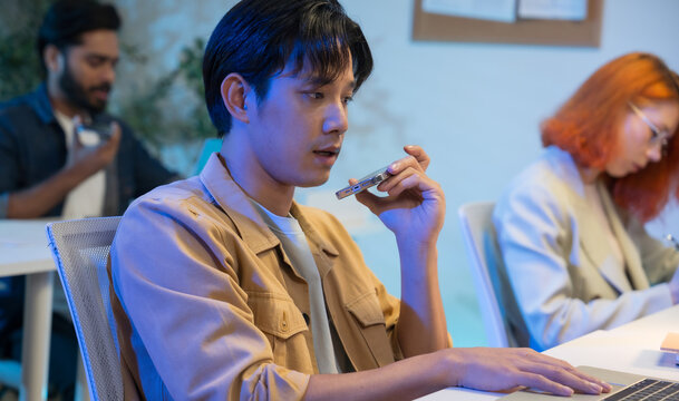 Asian man using AI voice assistant on smartphone in office, interacting with virtual assistant app for productivity, smart technology, and team collaboration.