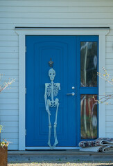 A skeleton figure stands at the vibrant blue door of a house, adding a playful touch to the entrance. The setting is bright with natural lighting, and the scene feels festive