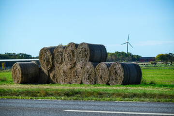 Hay bales are neatly stacked in a vibrant green field under blue skies. A wind turbine stands in the background, highlighting sustainable energy in the rural landscape
