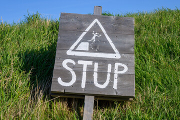 A warning sign stands on a grassy slope, alerting people to a steep drop ahead. The bright day adds clarity, emphasizing the caution needed while walking near the edge