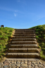 A series of stone steps ascends to a lush green hill under a bright blue sky. The area is peaceful, ideal for a quiet walk or relaxation