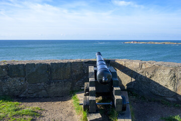 A historic cannon sits on a stone wall, pointing towards the vast ocean. Bright blue skies and gentle waves create a peaceful coastal scene with distant land visible