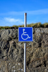 A blue handicap sign stands on a pole next to a sturdy stone wall. The sky is clear with a few clouds, indicating a sunny day. This area promotes accessibility for individuals