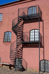 A steel spiral staircase winds upwards against a vibrant pink building. The scene captures a sunny day, highlighting the contrasting colors and unique architectural features