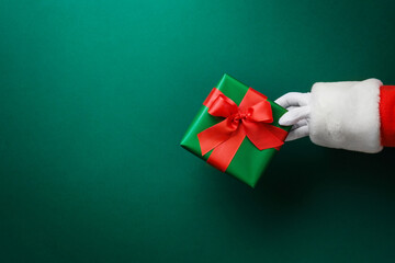 Santa Claus hand holding green gift box with red ribbon on festive green background with copy space for text, Christmas card design element.