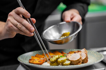A chef's hands are carefully plating a dish of pork tenderloin, roasted potatoes, and pickles using tweezers in a professional kitchen