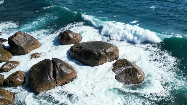 An awe-inspiring high-angle view captures the raw power and beauty of the ocean as vibrant turquoise and deep blue waves relentlessly crash against rugged, weathered coastal rocks. White foamy water c