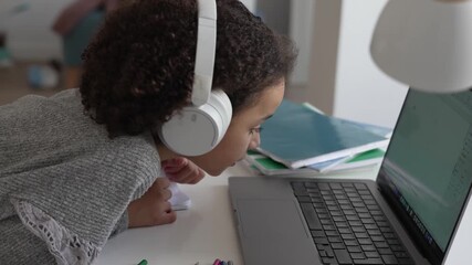 Primary school African American Student child with headphones hand writing in book using laptop. Distance learning online education. School girl studying at home with digital tablet computer and doing - Powered by Adobe