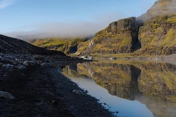 Icelandic landscape with volcanic sand and rocky cliffs reflected in lake.