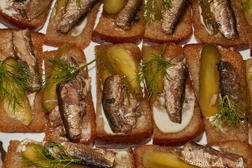 Close-up of delicious sandwiches with canned sprats, pickled cucumbers, and fresh herbs on toasted rye bread. Classic retro-style snack: sprats on crispy bread with mayonnaise and pickled cucumber.