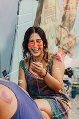 Mujer joven de cara pintada, con pintura en brazos y manos, sonriendo y sentada frente a un mural,...