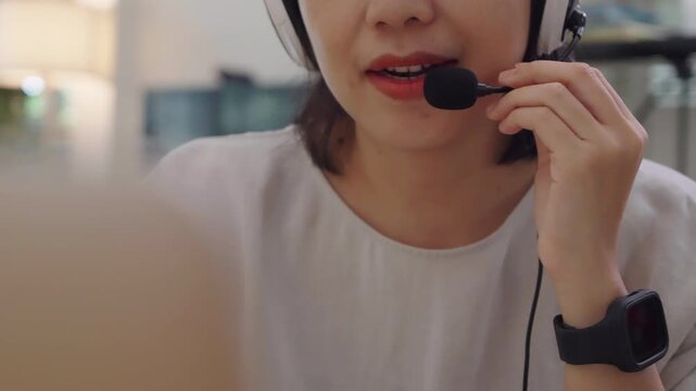Young Asian female agent chatting through a headset microphone at home, offering kind customer assistance and staying relaxed during remote work.