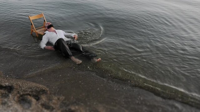 Conceptual scene of a man in wet clothes lying in shallow sea water near chair on beach, symbol of stress, burnout and emotional freedom outdoors.