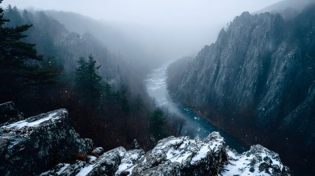 Dramatic winter mountain landscape with a winding river through steep misty cliffs blanketed in falling snow