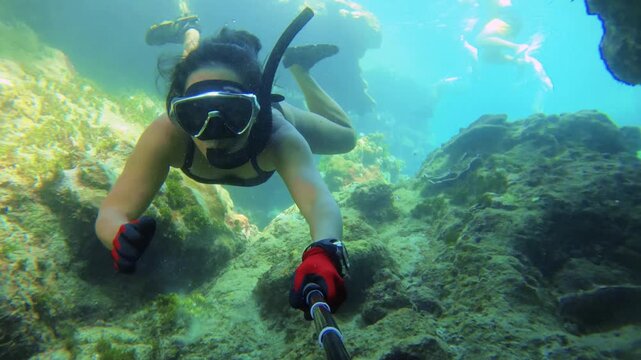selfie of an asian young female with tanned skin wearing bikini and mask snorkel in mouth free diving down into ocean water no fin kicking swimming around coral reef through narrow passages day time