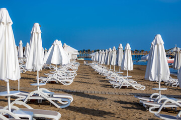 Rows of white sunbeds and closed umbrellas stand ready on an empty sandy beach, with turquoise sea and blue sky signaling the approaching summer season. Side, Antalya, Turkey.