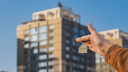 Hand Holding House Keychain on Background of Apartment Building