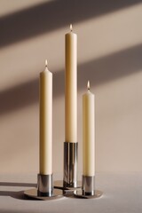 A still life composition of three ivory taper candles burning in sleek polished chrome holders with soft shadow play across a neutral beige surface background