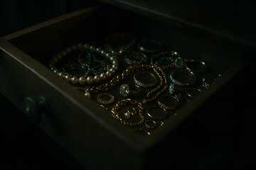 Hidden jewelry collection inside dark wooden drawer illuminated with dramatic low key lighting, luxury gold accessories and diamonds arranged in secret stash crime mystery atmosphere