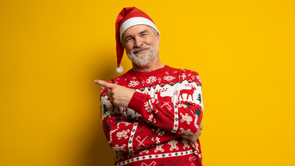 Merry mature man wear knitted christmas sweater Santa hat presenting something on yellow background