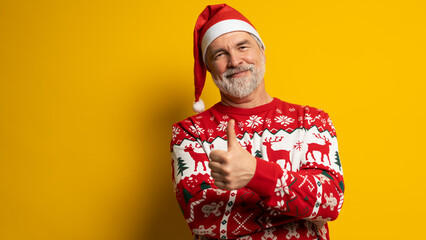 Merry mature man wear knitted christmas sweater Santa hat presenting something on yellow background