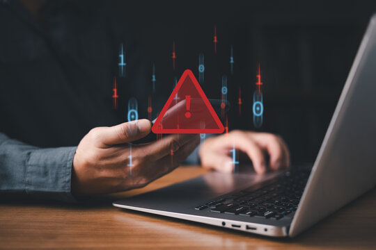 A person using a smartphone and laptop with a red warning alert icon, representing cybersecurity threat, malware attack, phishing, or data breach risk in the digital environment.