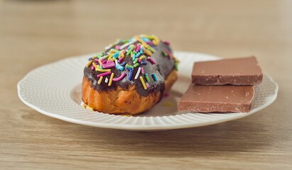 A delicious chocolate dessert on a white plate. A pastry, like an eclair cookie is covered in chocolate and sprinkles, next to squares of milk chocolate